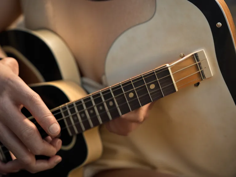 Mano de un adulto principiante formando el acorde de Sol con rasgueo básico en el mástil de una guitarra acústica.